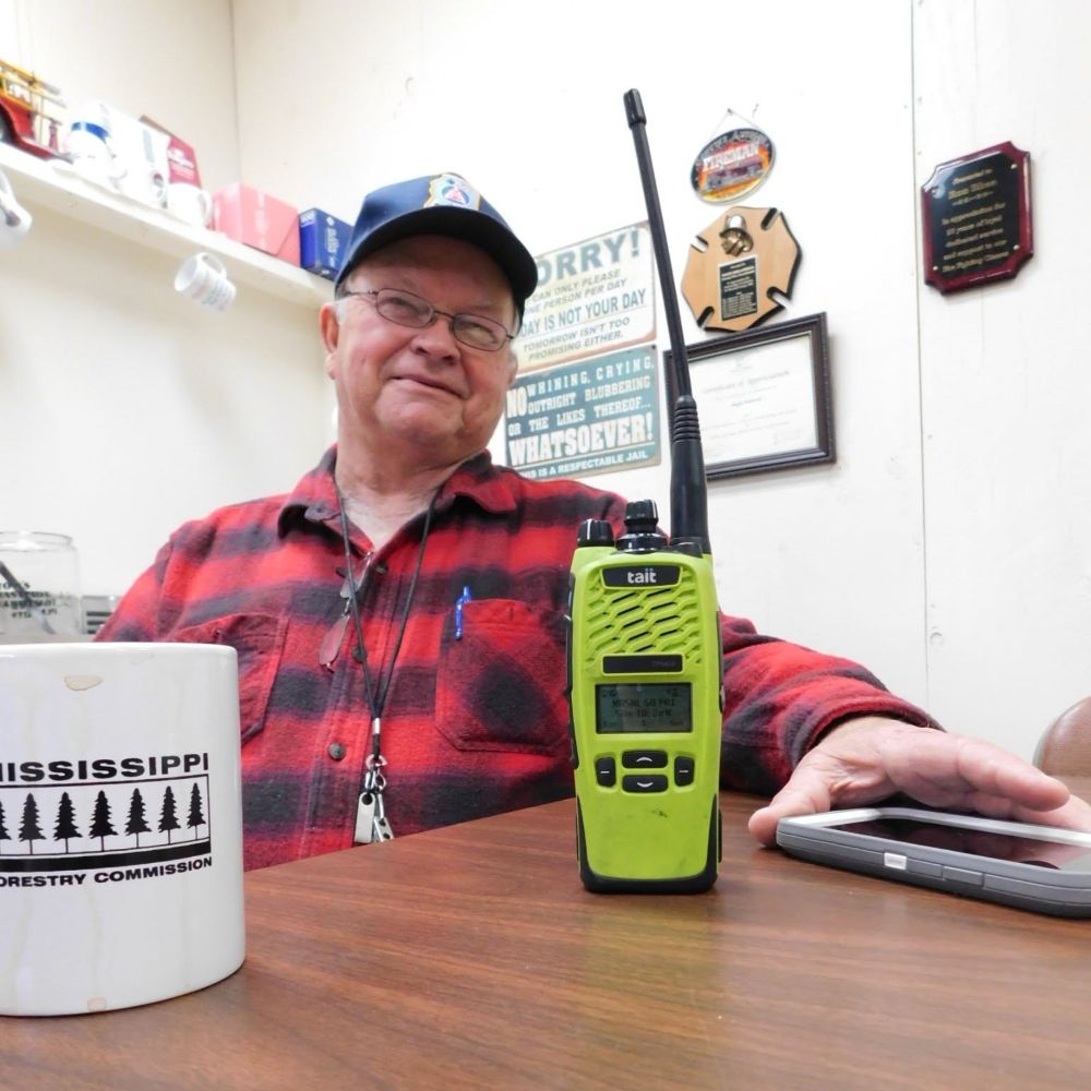 A man in glasses, a red and black plaid flannel shirt, and a baseball cap sitting at a desk. In the foreground, a high-visibility yellow Tait brand walkie-talkie stands upright next to a white coffee mug featuring the "Mississippi Forestry Commission" logo. A smartphone lies on the desk to the right. In the background, framed certificates, plaques, and a humorous sign hang on the wall.
